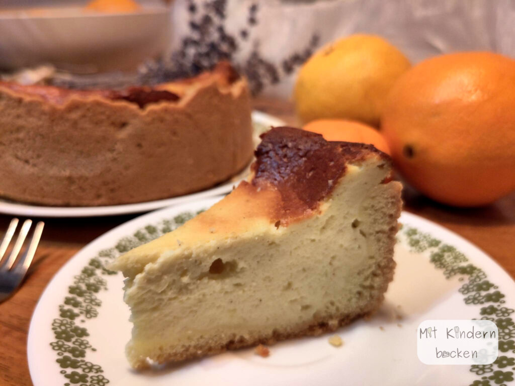 Ein Stück Käsekuchen auf einem Teller, im Hintergrund Käsekuchen, Lavendel und Orangen
